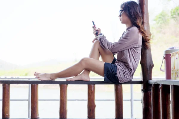 Mujer solitaria jugando al teléfono móvil mientras está sentado balcón de madera en el paseo marítimo. esta imagen para viajes, retrato y tecnología — Foto de Stock