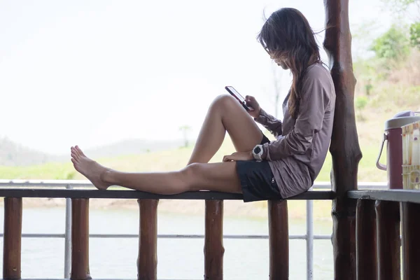 Mujer joven jugando al teléfono móvil mientras está sentado balcón de madera en el paseo marítimo. esta imagen para viajar, retrato y concepto de tecnología —  Fotos de Stock