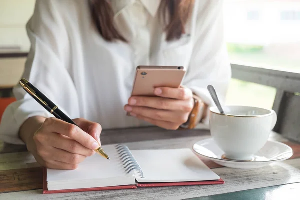 Wanita muda kemeja lengkap menggunakan ponsel untuk memeriksa sesuatu sambil memegang pena untuk menulis di notebook kosong dan memiliki cangkir kopi menempatkan di sampingnya. gambar ini untuk kemerosotan, pendidikan dan konsep potret — Stok Foto