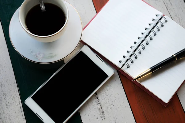 Pemandangan atas. Ponsel, cangkir kopi dengan kopi, pena ditaruh di atas kertas notebook. semua memakai kayu adalah latar belakang. gambar ini untuk bisnis, aksesori dan konsep pendidikan — Stok Foto