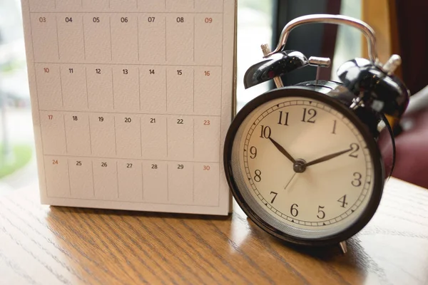 Reloj despertador colocado junto al calendario en la mesa de madera con ventana a —  Fotos de Stock