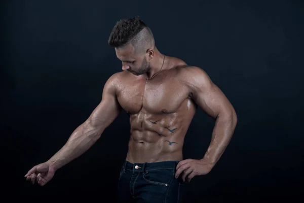 Jonge knappe gespierde sterke bodybuilder / fitness model met perfect lichaam kijkt naar zichzelf op een donkere achtergrond in een studio — Stockfoto