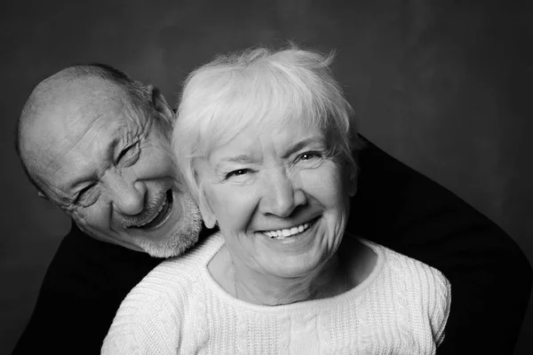 Black and white studio shot of handsome senior couple on a dark background. Man in black hugs his wife in white smiling and laughing looking at the camera. Love and devotion concept