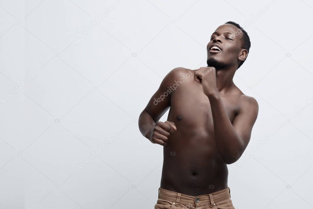 Handsome African American dark- skinned man is dancing enjoying of being himself and the beauty of his athletic body with eyes shut. Moving shirtless against gray studio wall