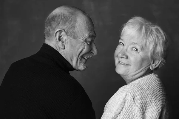 Retrato de amor sin fin. Retrato en blanco y negro de una feliz pareja de ancianos mirándose y sonriendo mientras están de pie en el interior mano a mano sobre un fondo oscuro — Foto de Stock