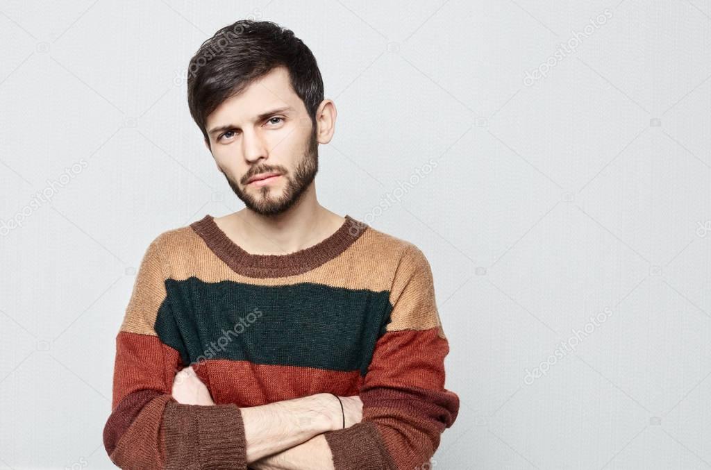 Studio portrait of young bearded hipster male looking upset, indifferent , shameful and Impolite. Dressed in old-fashioned colorful sweater guy crosses arms and stares in displeasure.