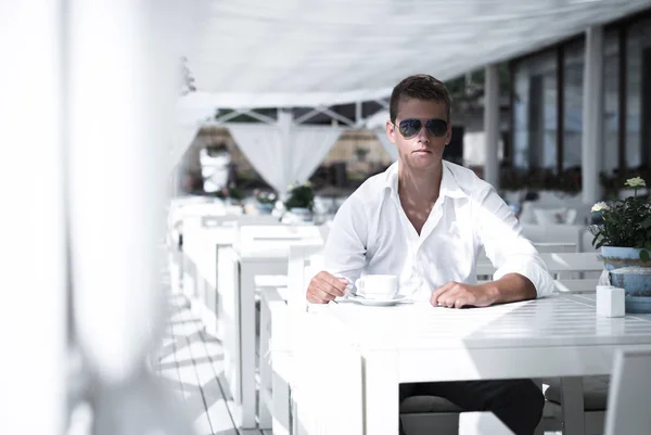 Молодий серйозний бізнес стиль чоловік п'є каву в ресторан на березі моря. Сидячи поодинці носіння стильний сонцезахисні окуляри і білу сорочку кавказьких хлопець дивиться на камеру. — стокове фото