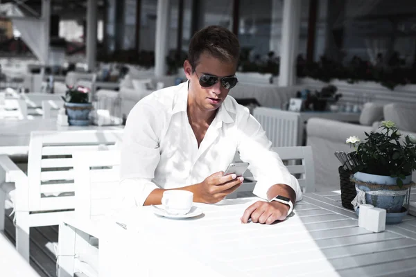 Молодий стильний і модний чоловік зі смарт-годинником на руці перевіряє телефон і п'є чай у барі під відкритим небом в сонячний літній день . — стокове фото