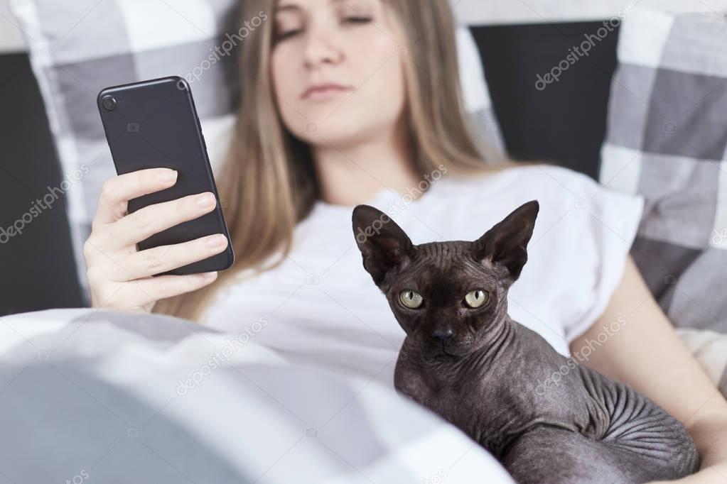 Portrait of sphinx cat looks forward at the camera and young woman behind with smartphone texting message in bed at home. Technology, pets, communication and people concept.