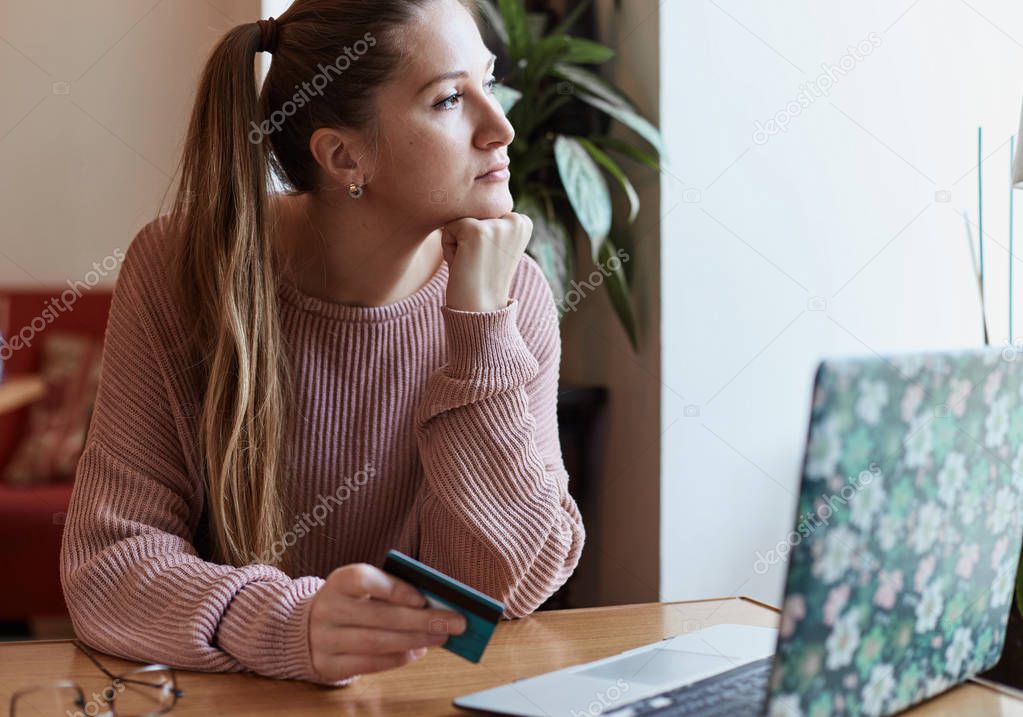 Modern technology and online payment.Young Caucasian beautiful lady is sitting at cafe table with her laptop opened, holding credit card, looking at the window trying to make an important decision.
