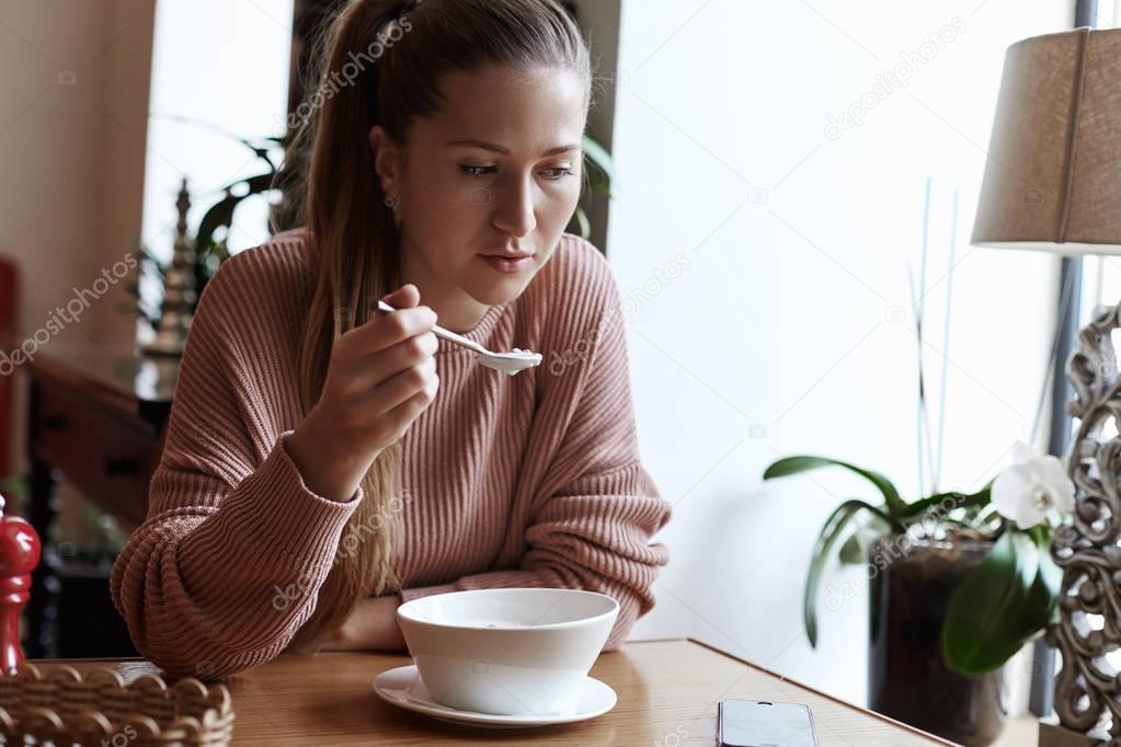 Healthy eating, balanced diet, food and people concept. Young Caucasian attractive lady is having her breakfast bowl with granola and farm yogurt in organic modern cafe. Beautiful girl is looking at her phone, waiting for message while her eating .