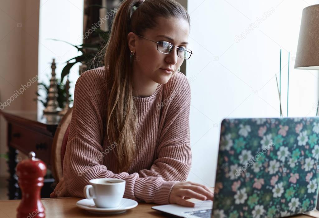 Close up shot of young attractive Caucasian student girl with long hair wearing eyeglasses, sitting at cafeteria with fancy laptop. Freelance worker is doing her job drinking coffee during daytime.