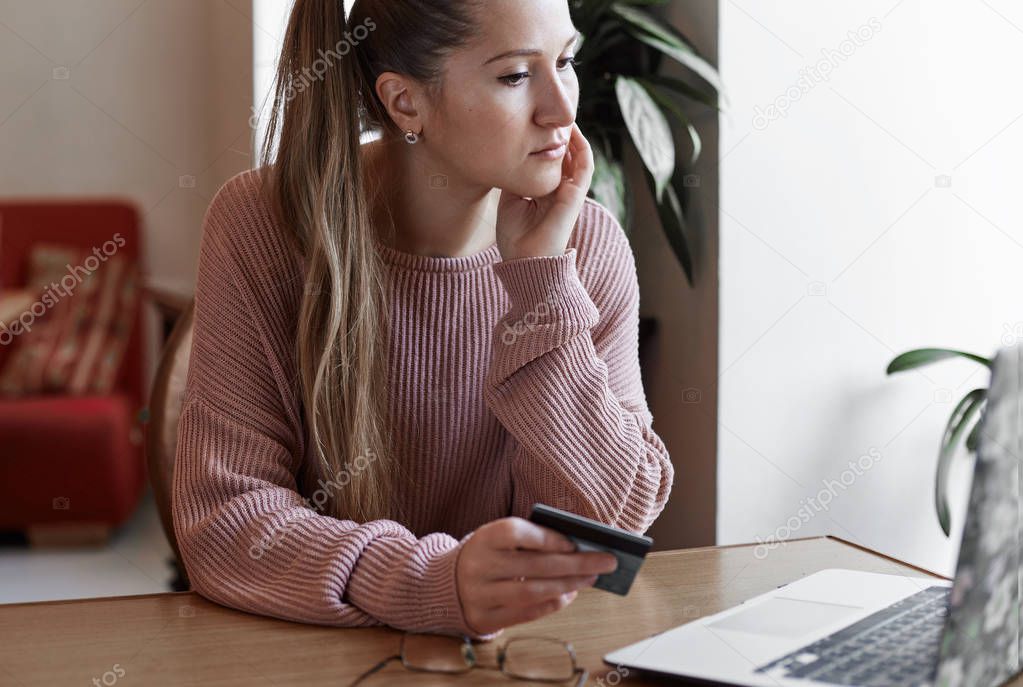 Internet banking, e-commerce and online trading concept. Close up shot of  young beautiful Caucasian lady holding credit card in her hands and thoughtful looking at the screen of her modern laptop while paying bill at cafe.