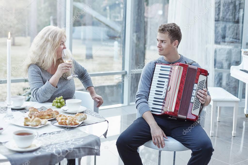 Family moments at home. Young handsome male is playing accordion for his senior beautiful mother. Attractive mature Caucasian woman is looking at her adorable son with love drinking white wine during lunch time at stylish luxury house.