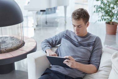 Çekici genç kafkas erkek evde tablet, işten sonra beyaz koltukta tek başına oturan ile rahatlatıcı. Genç çekici şık adam online app onun modern gadget'ı kullanarak kız arkadaşı ile görüntülü görüşme.