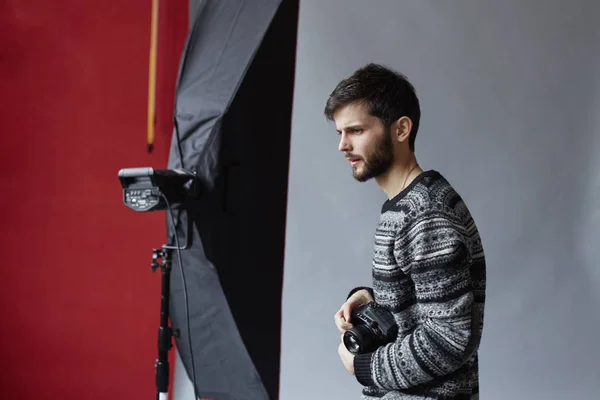 Studio portrait of handsome Caucasian man posing on grey background. Young attractive bearded photographer with camera in his hands is watching pictures.