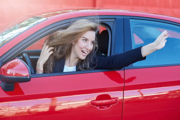 Portret van jonge en mooie Kaukasische business dame met lang haar dragen van formele pak op het wiel van rode moderne auto opknoping en flirten met haar college tijdens het parkeren bij het Bureau. — Stockfoto