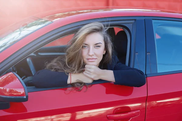 Portrait of young attractive Caucasian business lady with long hair wearing formal suit posing on the wheel of red modern car smiling gently and looking deeply to the camera with beautiful blue eyes. — Stock Photo, Image