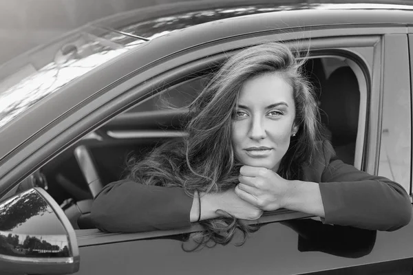 Retrato en blanco y negro de la joven atractiva dama de negocios caucásica con el pelo largo usando traje formal posando en la rueda del coche moderno rojo sonriendo suavemente y mirando profundamente con hermosos ojos . — Foto de Stock