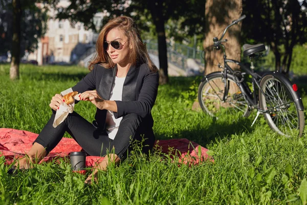 Persone, cibo e stile di vita. Colpo all'aperto di attraente giovane manager donna vestita di nero abito alla moda e occhiali provenienti in bicicletta per fare colazione nel parco prima del suo ufficio stelle del lavoro . — Foto Stock