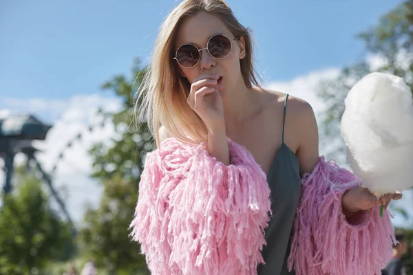 Primer plano retrato de mujer hipster rubia feliz con gafas de sol, vestido verde y abrigo brillante comer azúcar de algodón blanco al aire libre en el parque de atracciones. Juventud, viajes, aventura, concepto de vacaciones . — Foto de Stock