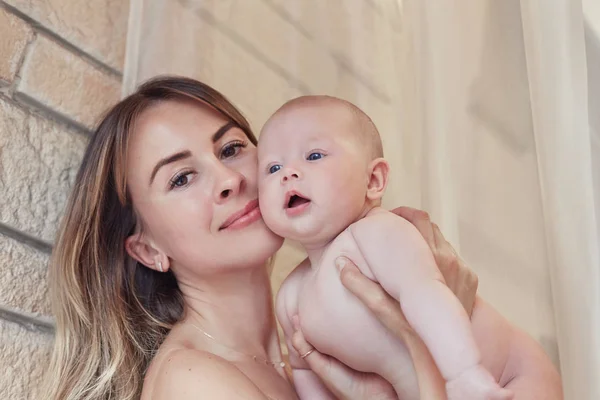 Giovane madre che guarda la macchina fotografica con il suo piccolo bambino dagli occhi azzurri che non indossa nulla di rilassante e gioca in camera da letto durante il fine settimana insieme, mattina pigra, scena calda e accogliente. Mattone muro di sfondo . — Foto Stock