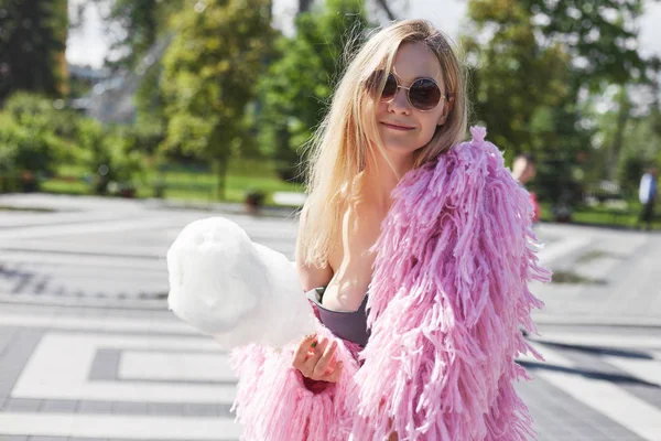 Chica estudiante rubia caucásica con algodón de azúcar en la mano con abrigo funky rosa y gafas de sol redondas descansando en el parque de la ciudad después de conferencias en la universidad, mirando a la cámara con sonrisa de ensueño alegre . —  Fotos de Stock