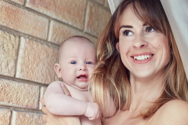 Retrato familiar de la sonriente madre moderna y su bebé sobre fondo de pared de ladrillo. Emocional toma de hermosa dama europea sonriendo y divirtiéndose con su hijo mirando con miedo a la cámara — Foto de Stock