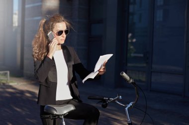 Çevre dostu kadın Yöneticisi resmi takım elbise ve ciddi bir yüz ifadesi ile rapor okuma güneş gözlüğü. Şık ve çekici Bayan Onun Bisiklet bir telefon görüşmesi yapmak için yolda ofisine durur..