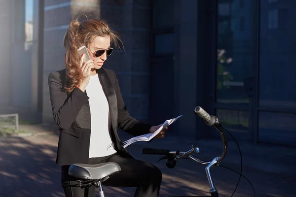 Environmentally friendly female manager in formal suit and sunglasses reading report with serious face expression. Stylish and attractive lady stops her bicycle on the way to office to make a phone call.