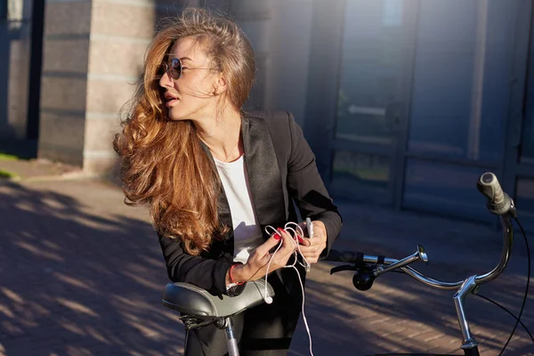 Environmentally friendly female manager in formal suit and sunglasses dance outdoors. Stylish and attractive cheerful lady stops her bicycle on the way to office to turn on music player on her phone.