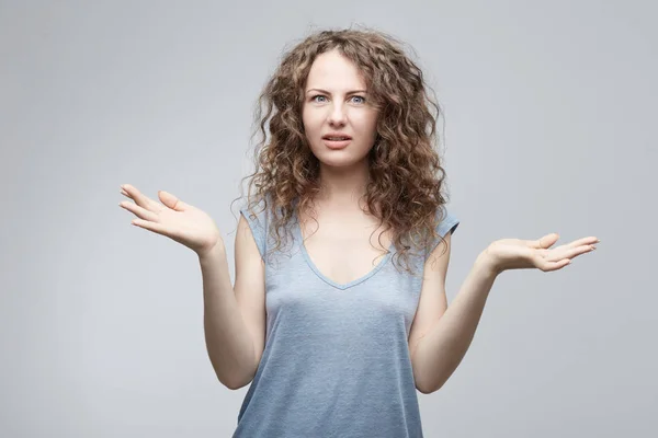 Mujer europea dudosa confusa y confusa con el pelo largo rizado en una camiseta gris casual encogiéndose de hombros en un gesto cuestionable de incertidumbre y manteniendo la boca abierta, habiendo confundido la mirada despistada . — Foto de Stock