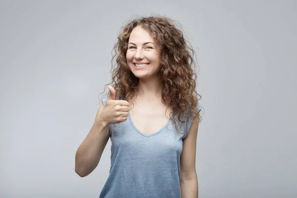 Retrato de una joven mujer caucásica feliz dando en camisa casual gris mostrando el pulgar hacia arriba o como gesto, mirando y sonriendo cámara con expresión feliz. Emociones humanas, expresiones faciales . —  Fotos de Stock