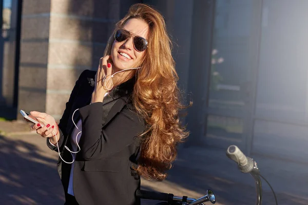 Gerente femenina respetuosa con el medio ambiente en traje formal y gafas de sol bailan al aire libre. Elegante y atractiva dama alegre detiene su bicicleta en el camino a la oficina para encender el reproductor de música en su teléfono . —  Fotos de Stock