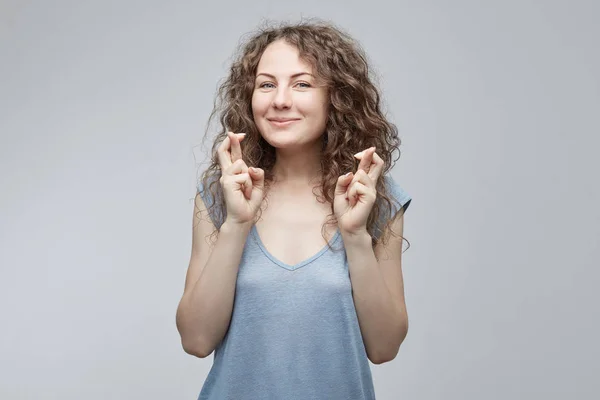 Carino femmina dagli occhi azzurri caucasici con bel viso in t-shirt grigia incrociando le dita per buona fortuna, sperando che tutti i desideri si avverino, avendo eccitato l'occhiata. Emozioni e sentimenti umani — Foto Stock