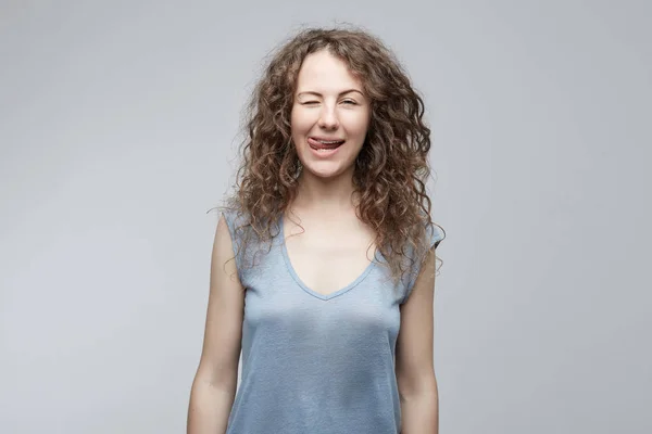 Foto de estudio de atractiva chica divertida optimista con el pelo rizado mostrando su lengua y ojos parpadeantes mientras posando sobre fondo blanco. Emocional dama caucásica haciendo mueca . — Foto de Stock