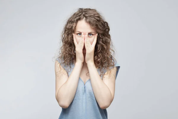 Mujer caucásica joven preocupada con el pelo rizado cubriendo la cara con ambas manos sintiéndose estresada, mirando a la cámara a través de los dedos con grandes ojos azules. Disparo aislado sobre fondo gris . — Foto de Stock