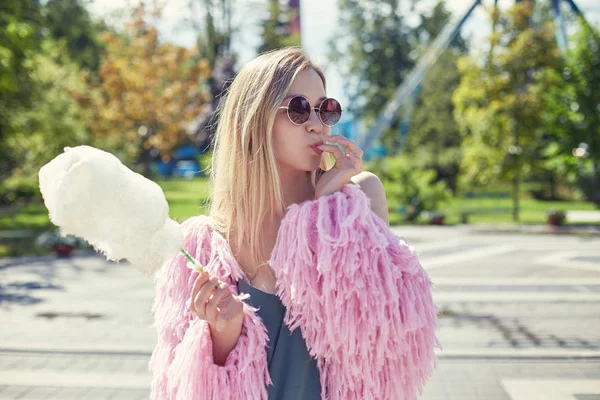 Joven bastante de moda adolescente rubia hembra se está divirtiendo en el parque de atracciones disfrutando de sus vacaciones y dulce algodón de caramelo. Mujer caucásica descansando afuera en el parque de la ciudad usando chaqueta extravagante rosa . —  Fotos de Stock