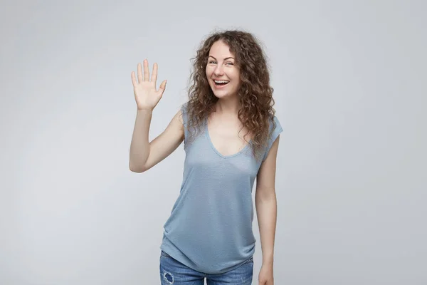 Vriendelijk ogende beleefd jonge Kaukasische vrouw gekleed in een grijs t-shirt zeggen hi, zwaaien van haar hand. Positieve menselijke emoties, gevoelens, gezichtsuitdrukkingen, houding en reactie. — Stockfoto