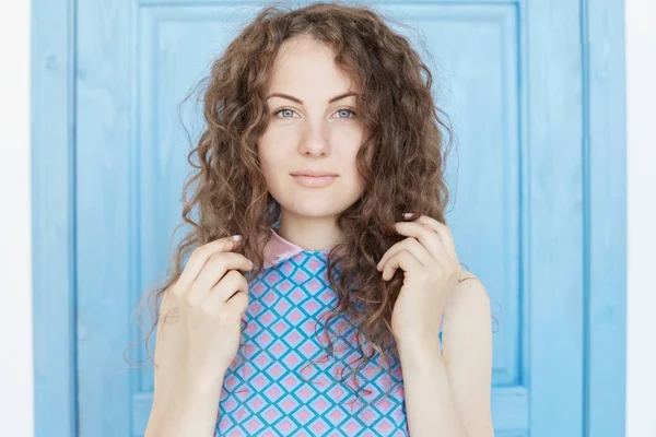 Close up visão isolada da bela jovem morena com pele saudável perfeita e olhos azuis, olhando em linha reta com expressão facial sedutora pensativa, tocando o cabelo encaracolado, fundo azul . — Fotografia de Stock