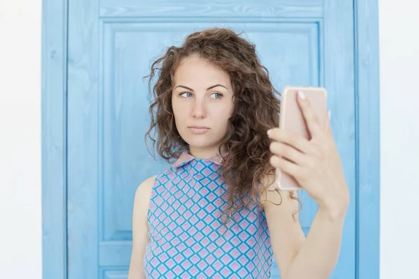 若い美しい白人巻き毛髪女性彼女の手でスマート フォンのデジタル カメラを持ってして、selfie のスタジオ撮影をクローズ アップ青い背景に立って自分のセルフ ポートレート. — ストック写真