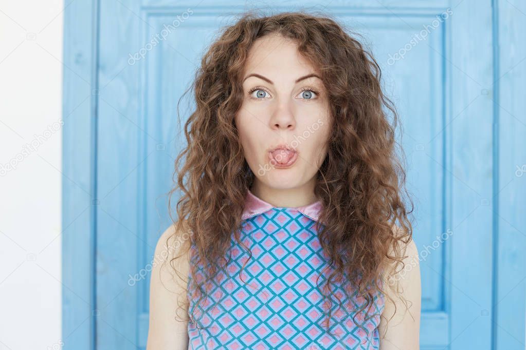 Young pretty fashionable lady with curly hair sticking out tongue having funny look isolated over door background. Blue eyed student girl having fun being happy to finish her studying successfully