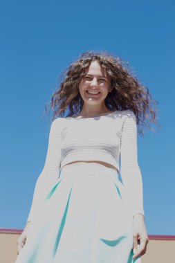 Dikey beyaz üst ve mavi etek giyen, güzel havanın tadını, güneşli günde şehirde yürüyüş kıvırcık saçlı genç çekici kadın portresi, kamera gözlü gülüyorsun zevk kapalı.