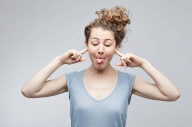 Komik, duygusal, genç beyaz kız öğrencinin yapması, ağız yapma, onun dilini ve bir palyaço veya küçük yaramaz çocuk gibi davranan biri alay kulakları kapanış gri t-shirt giyen.