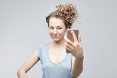 Beyaz stüdyo duvar arka planı poz kameraya gülümseyen genç beyaz neşeli bir kadın gri t-shirt yapımı selfie modern cep telefonu kullaranarak topuz kıvırcık saçlı. Teknoloji kavramı.