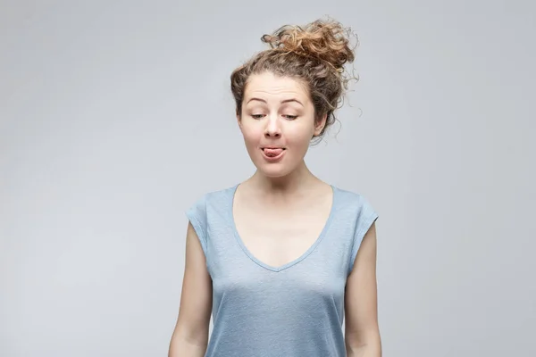 Divertida, emocional y joven estudiante caucásica vistiendo una camiseta gris haciendo muecas, haciendo bocas, sacando la lengua tratando de llegar a su nariz molestando a alguien, actuando como una niña traviesa . — Foto de Stock