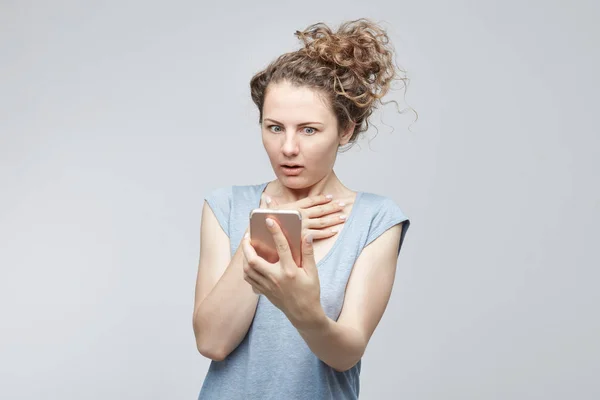 Retrato de estudio de una joven dama caucásica que parece asustada y confundida leyendo un mensaje de texto o un correo electrónico sosteniendo un teléfono móvil habilitado para la web con mal funcionamiento, con aterrorizada expresión de la cara preocupante . —  Fotos de Stock