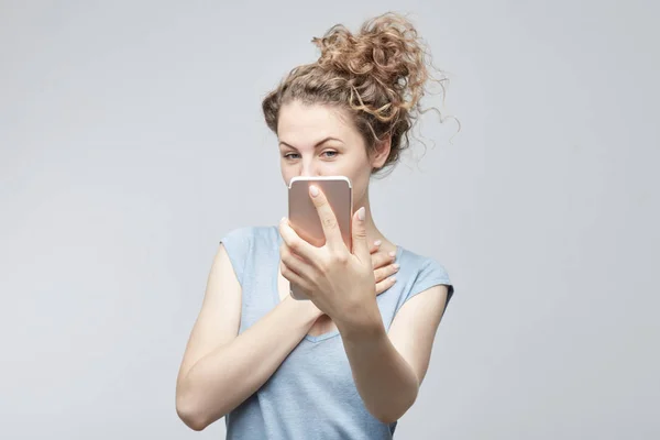 Selektiver Fokus auf das Telefon. Dame mit lockigem Haar im Dutt in grauem T-Shirt macht Selfie mit modernem Handy, blickt kokett in die Kamera, posiert vor weißem Studiowand-Hintergrund. Technologiekonzept. — Stockfoto