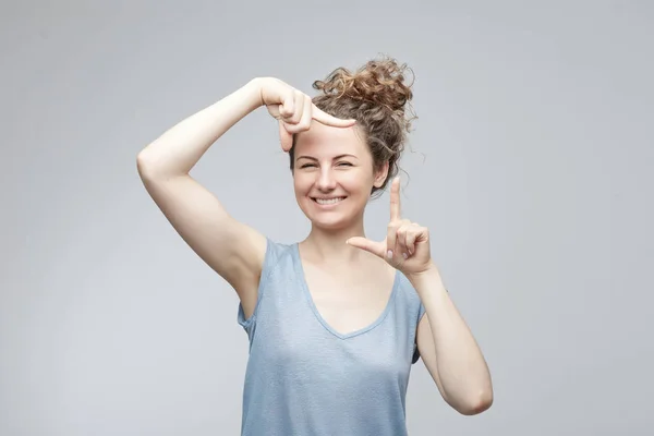 Gebaren vinger frame. Portret van aantrekkelijke vrolijke mooie jonge Kaukasische vrouw in casual t-shirt, glimlachend op camera & gebaren van de vinger frame, permanent tegen geïsoleerde grijze achtergrond binnenshuis. — Stockfoto