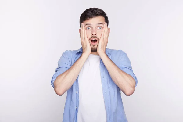 Sorprendido joven guapo habiendo abierto los ojos y la boca cogido de las manos en las mejillas mirando con asombro y sorpresa. Varón emocional siendo sorprendido después de escuchar noticias de terror. Concepto de personas y emociones — Foto de Stock